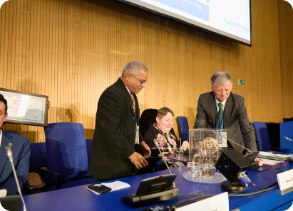 Officials and delegates participating in a formal event, standing around a podium with microphones and documents.