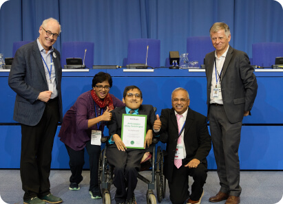 A group of five people posing together at a conference, with one person in a wheelchair holding a certificate and smiling, while others stand beside showing support.