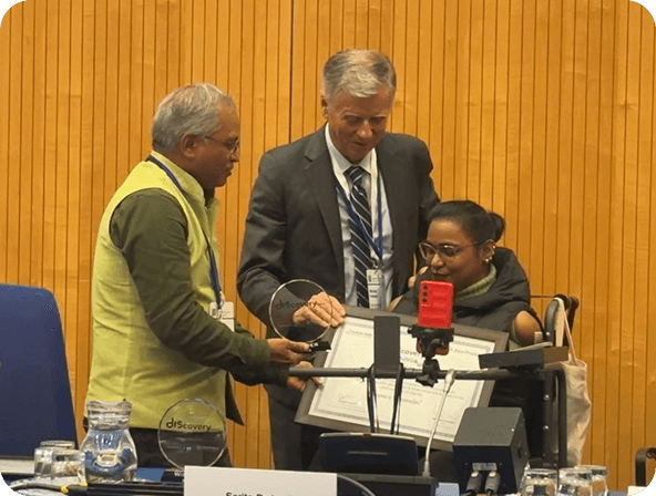 Three individuals at a formal event smiling and holding certificates and trophies, standing together in front of a wooden panel backdrop during an award presentation.