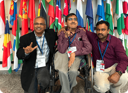 Three men with conference badges are smiling and posing together in front of a display of international flags at the Zero Project event.